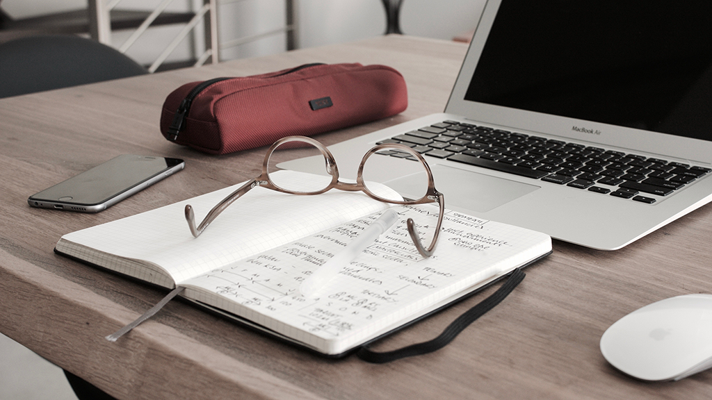 Brille und Notizbuch neben dem Laptop, als Zeichen für genaue Recherche.